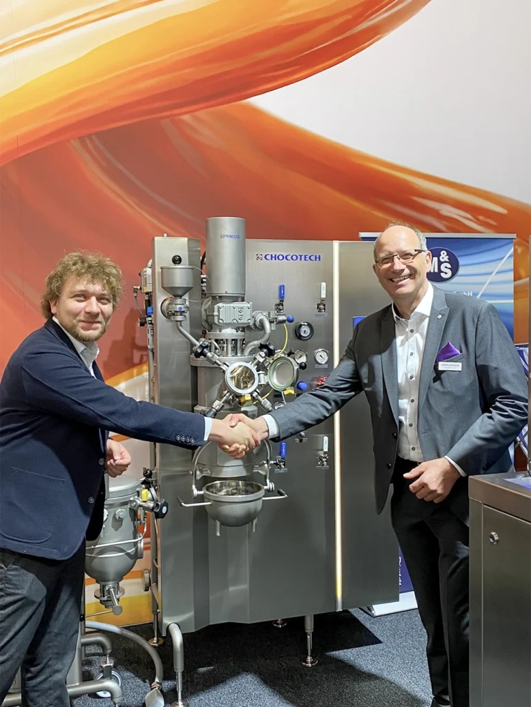 Chocotech GmbH, Theobald Huch Handschlag mit Timo Heinen Heinen von Industriedesign GmbH, vor dem neuen Satzkochers, der PRINCESS Candy präsentiert auf der Peitzmeier Maschinenbau GmbH präsentiert den neuen OMNI-GRID TURN Schleifer welcher von Heinen Industriedesign Gestaltet wurde auf der Interpack 2026 in Düsseldorf